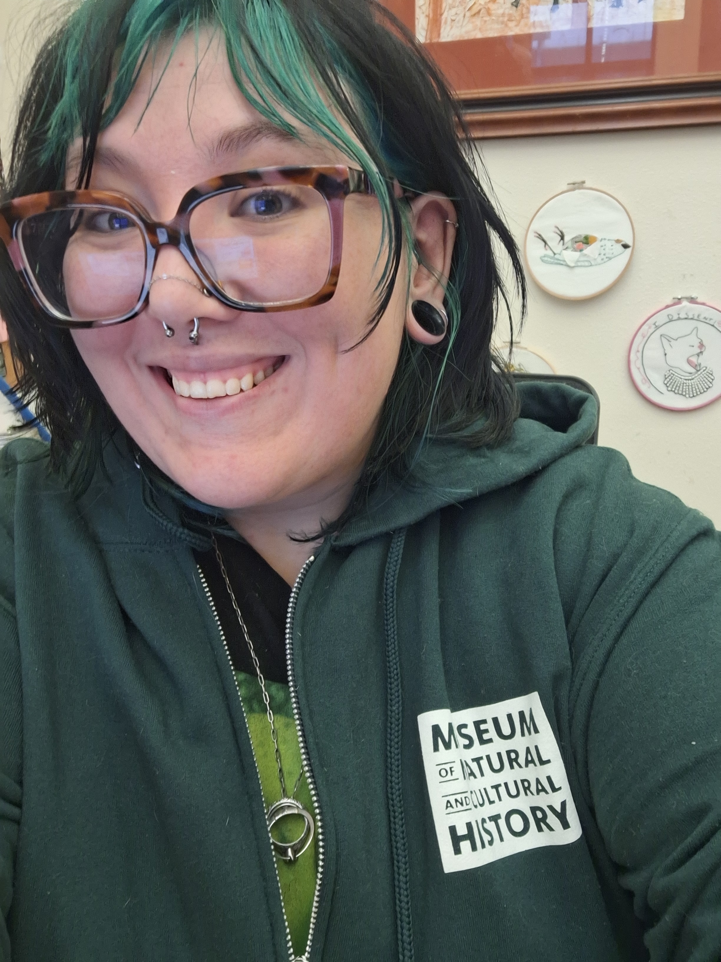 Will Coultley sits facing the camera with a smile. Their hair is black and green and they are wearing a dark green sweater with the logo for the University of Oregon's Museum of Natural and Cultural History